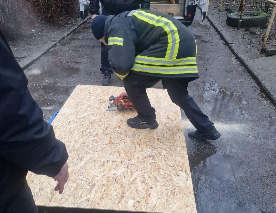 Спеціаліст у захисній формі зі світловідбивальними смугами розпилює лист плити ОСБ за допомогою електричної дискової пилки. Робота ведеться на вулиці на фоні вологого асфальту.