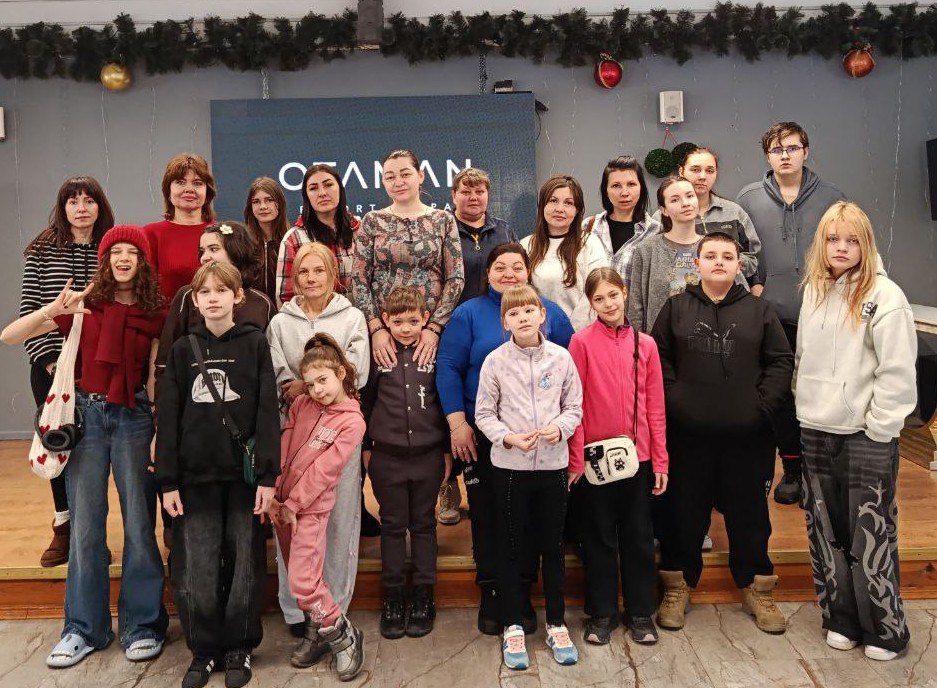 Групове фото дорослих та дітей у приміщенні на фоні стіни з логотипом. Зал прикрашений новорічними гірляндами та кулями.