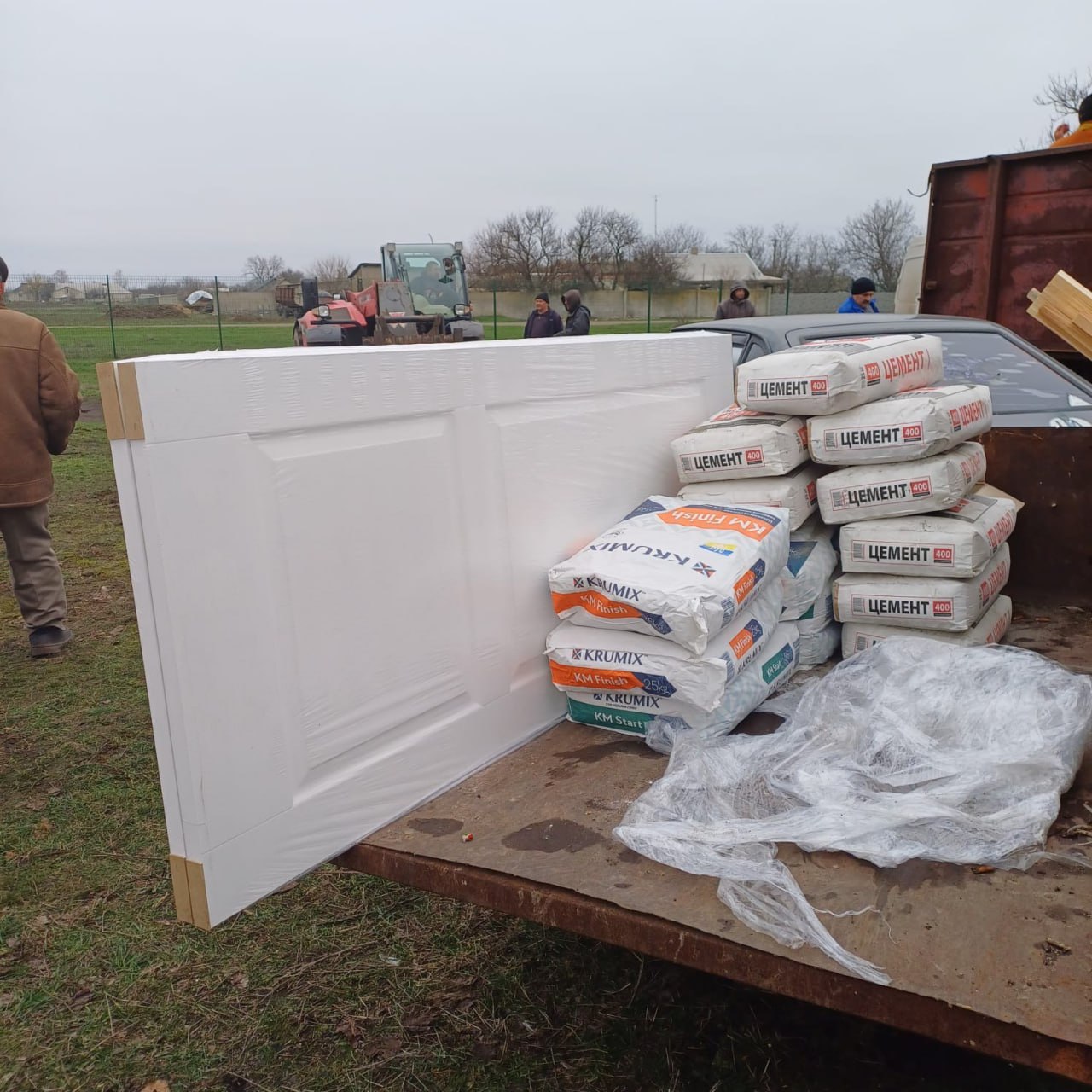 На відкритій платформі вантажівки лежать складені будівельні матеріали: білі міжкімнатні двері в захисній плівці та стоси мішків із цементом і шпаклівкою. На задньому плані, на відкритій місцевості, видно людей та техніку, що задіяні у процесі розвантаження.