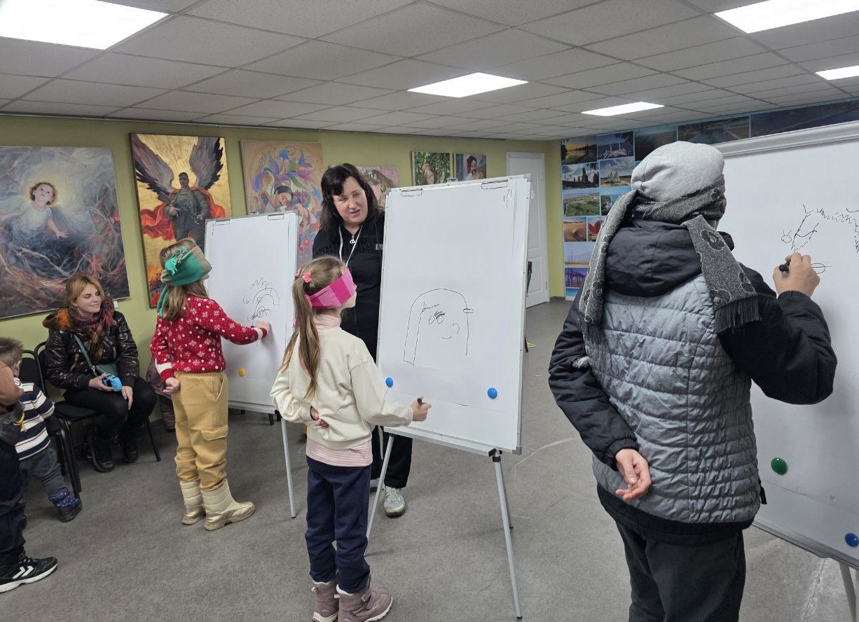 Діти та підлітки беруть участь у творчому конкурсі в приміщенні хабу. Учасники із зав'язаними очима намагаються малювати маркерами на білих фліпчартах під наглядом дорослої жінки. На стінах кімнати розвішані картини та численні фотографії.