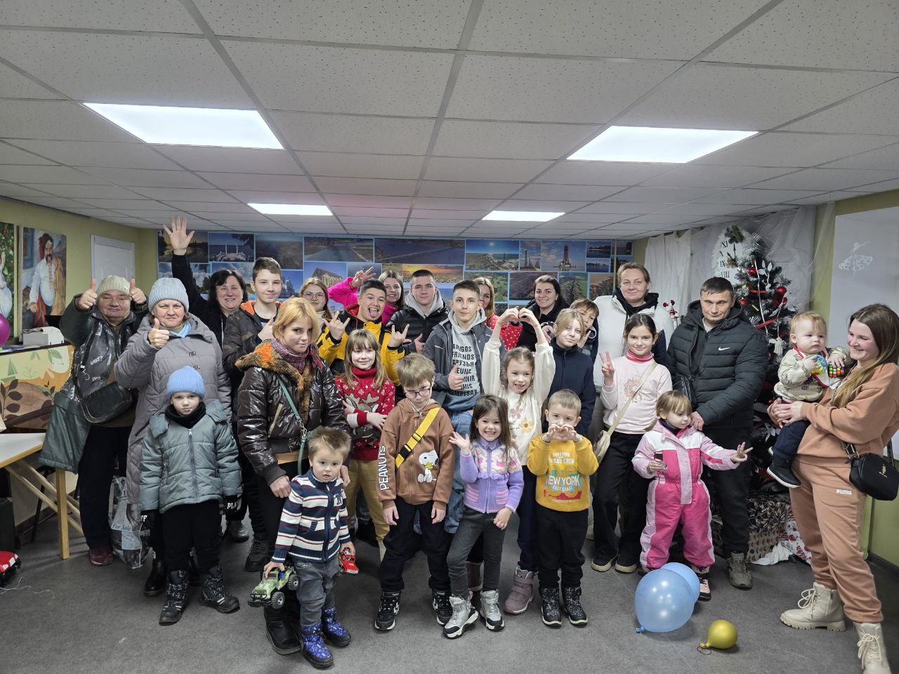 Велика група людей, серед яких дорослі, підлітки та маленькі діти, позують для спільного фото в приміщенні. Дехто з учасників посміхається, показує жест «клас» або складає руки у формі серця. На задньому плані видно стіну з численними фотографіями та прикрашену ялинку.