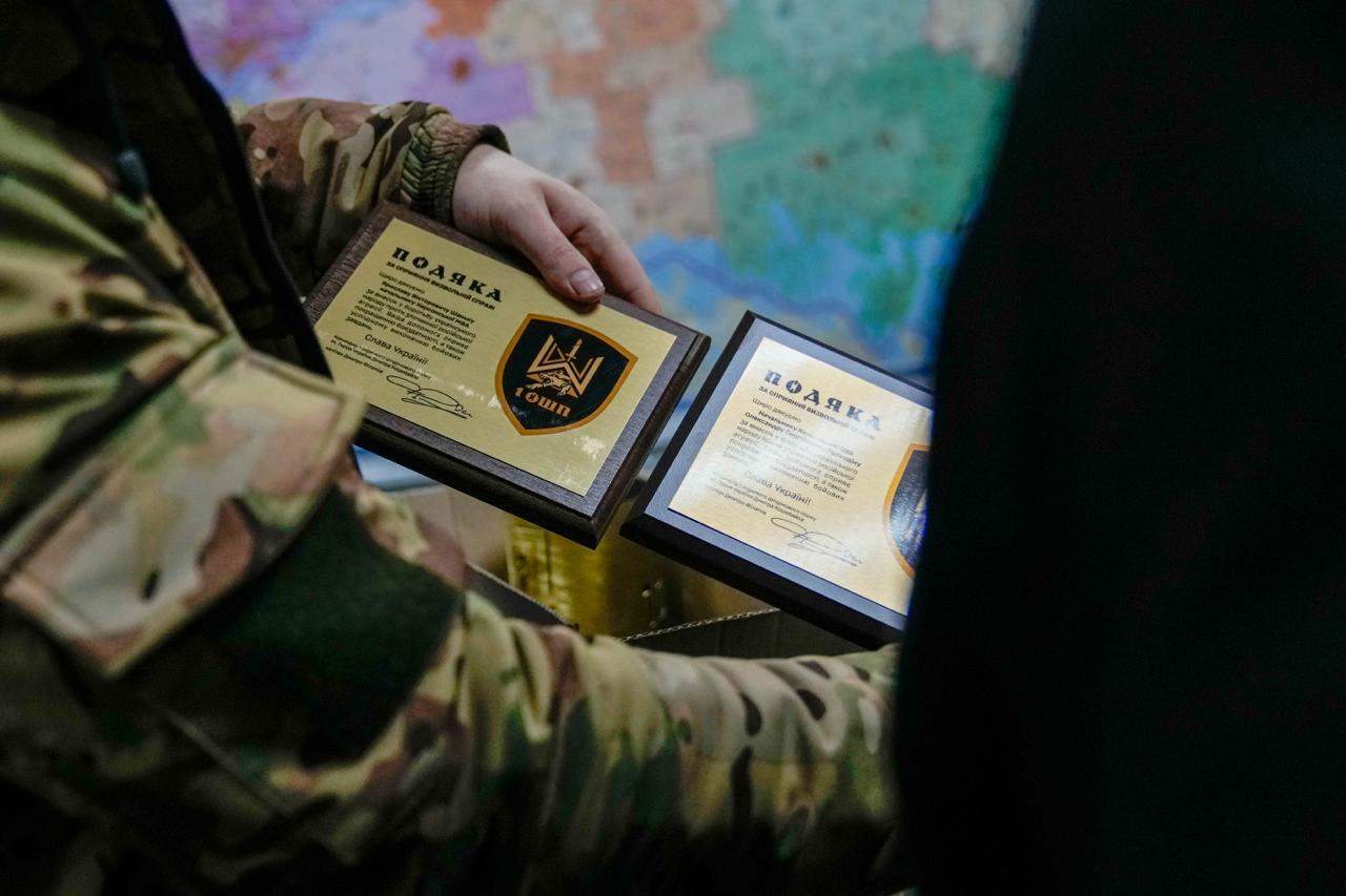Людина у камуфляжній формі тримає дві нагородні плакетки з написом «Подяка». На плакетках зображено герб підрозділу «1 ОШП» та текст із висловом вдячності за сприяння визвольній справі. Фоном слугує розмита карта.
