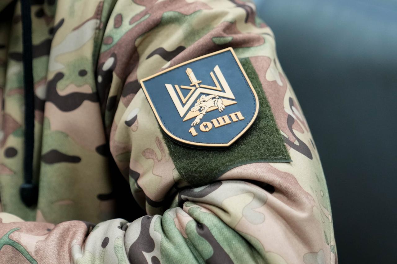Крупний план плеча військовослужбовця у камуфляжній формі із закріпленим шевроном. На шевроні темно-синього кольору зображено золотистий герб із вовком, мечем та написом «1 ОШП».