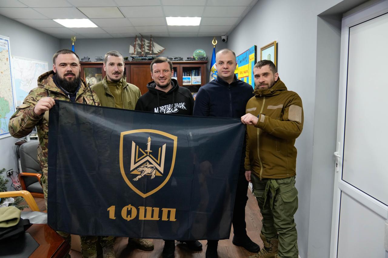 Група з п'яти чоловіків у кабінеті тримає великий темний прапор із золотистим гербом підрозділу «1 ОШБ». Деякі чоловіки одягнені у військову форму, інші — у цивільний одяг. На фоні видно офісні меблі, карту на стіні та макет корабля.