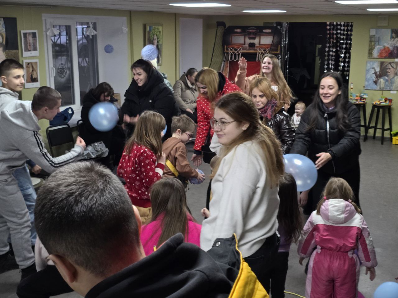 Група дорослих та дітей активно проводить час у просторому приміщенні хабу. Учасники граються з блакитними повітряними кульками, рухаючись по кімнаті. На задньому плані видно зону відпочинку з кріслами-мішками та ігровий автомат для баскетболу.