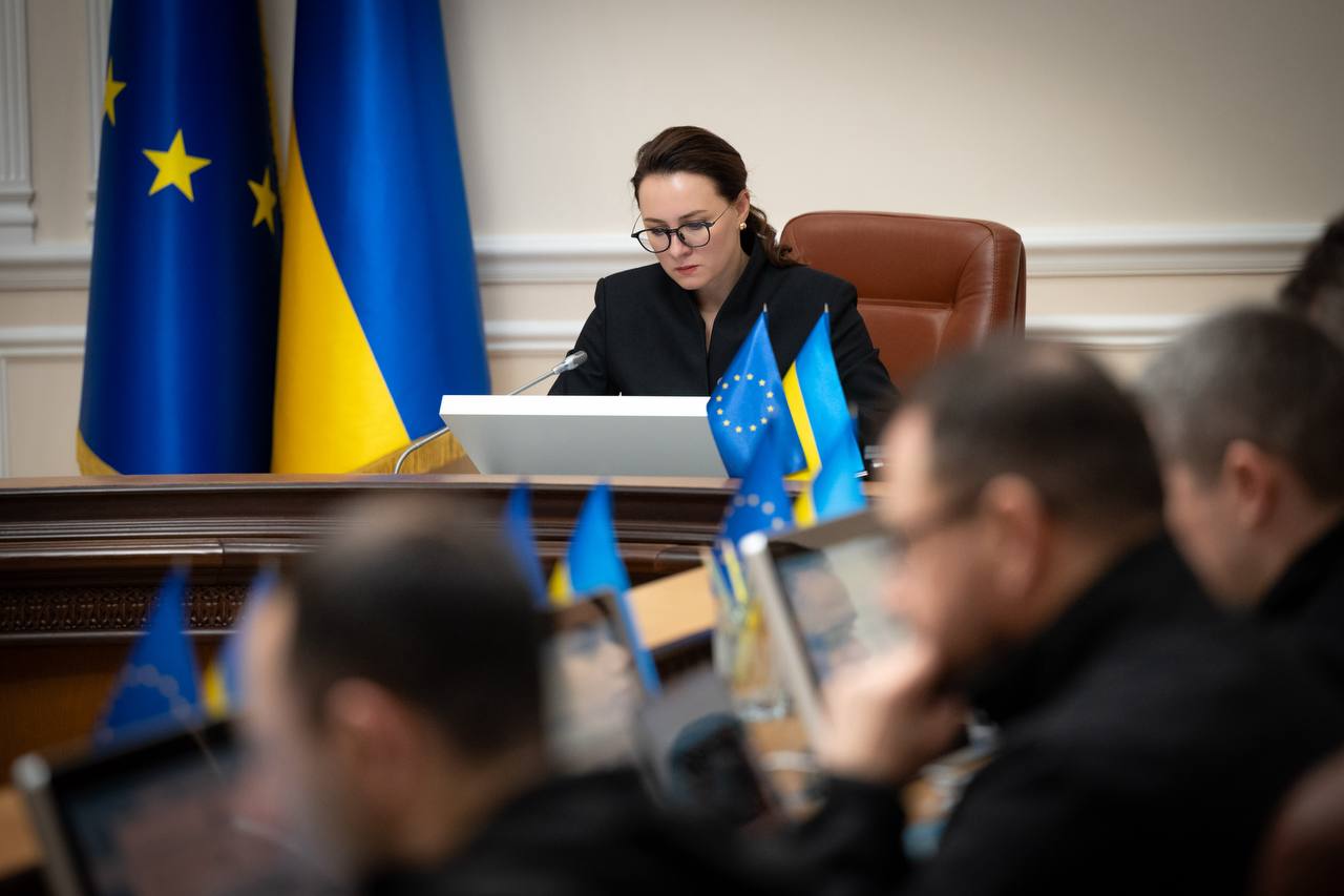 Жінка в окулярах та офіційному одязі сидить за робочим столом у залі засідань і зосереджено працює за монітором. На фоні та перед нею встановлено прапори України та Європейського Союзу. На передньому плані видно розмиті силуети інших учасників зустрічі.