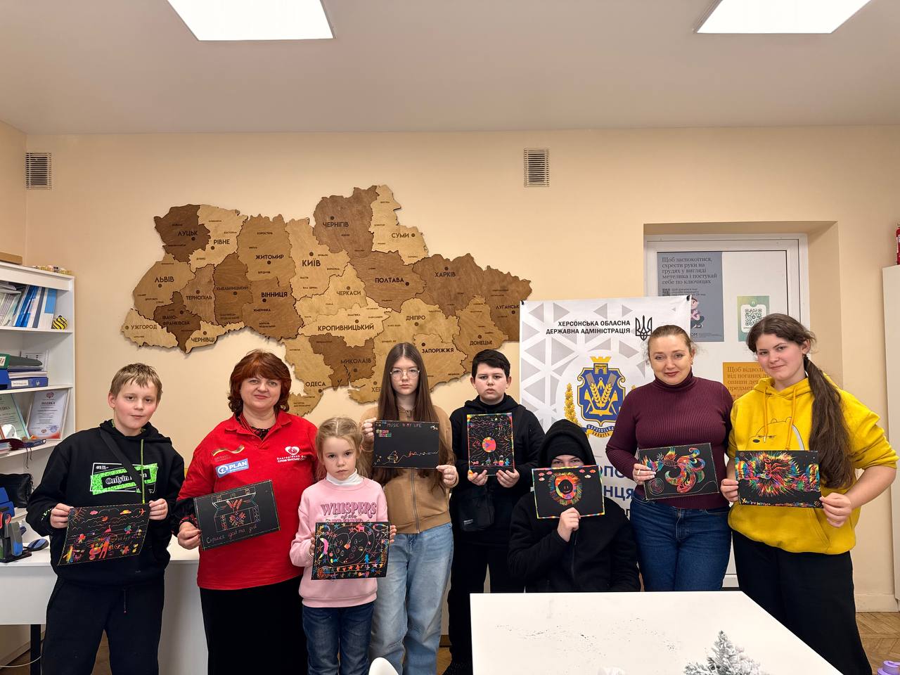 Групове фото дітей та дорослих у «Центрі допомоги херсонцям». Учасники стоять у світлій кімнаті на фоні великої дерев’яної мапи України та тримають у руках свої яскраві малюнки, виконані в техніці граттаж.