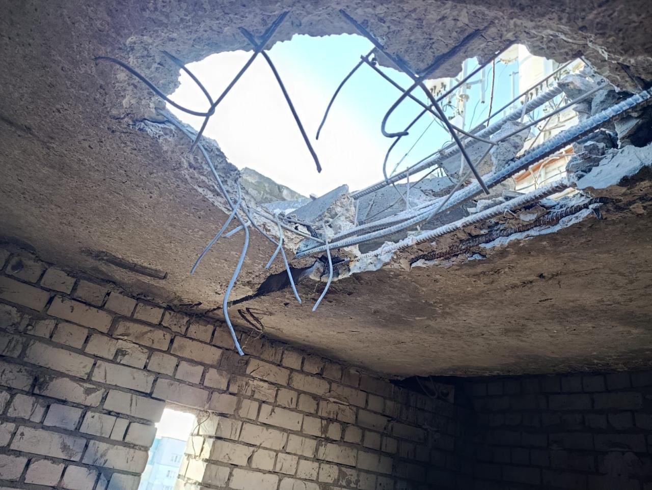 Наскрізний отвір у пошкодженій бетонній стелі будівлі, крізь який видно небо. З розлому виступає деформована металева арматура, а стіни приміщення викладені з білої цегли.