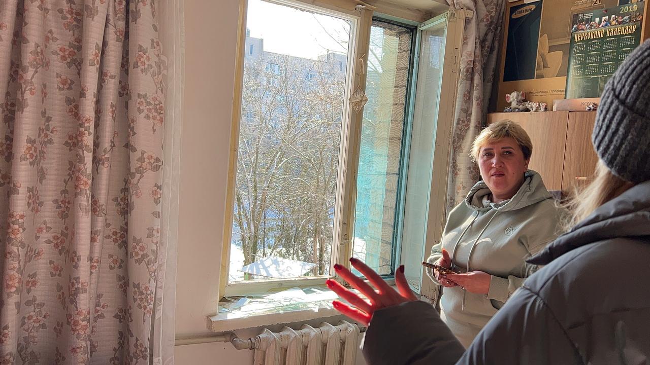 Жінка стоїть у кімнаті біля вікна, в якому вибите скло. На підвіконні лежать уламки, вікно відчинене, а за ним видно засніжені дерева та іншу будівлю. У кімнаті також помітні штори з квітковим принтом та меблі з календарем.