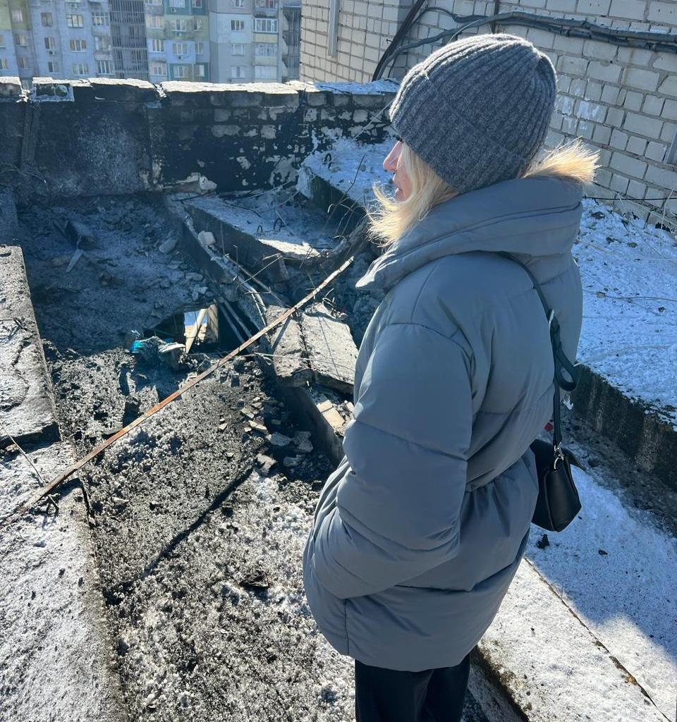 Жінка в зимовому одязі стоїть на даху будівлі поруч із місцем влучання. Видно значні руйнування бетонних конструкцій, вирву, розкидане будівельне сміття та сніг. На фоні — міська забудова.