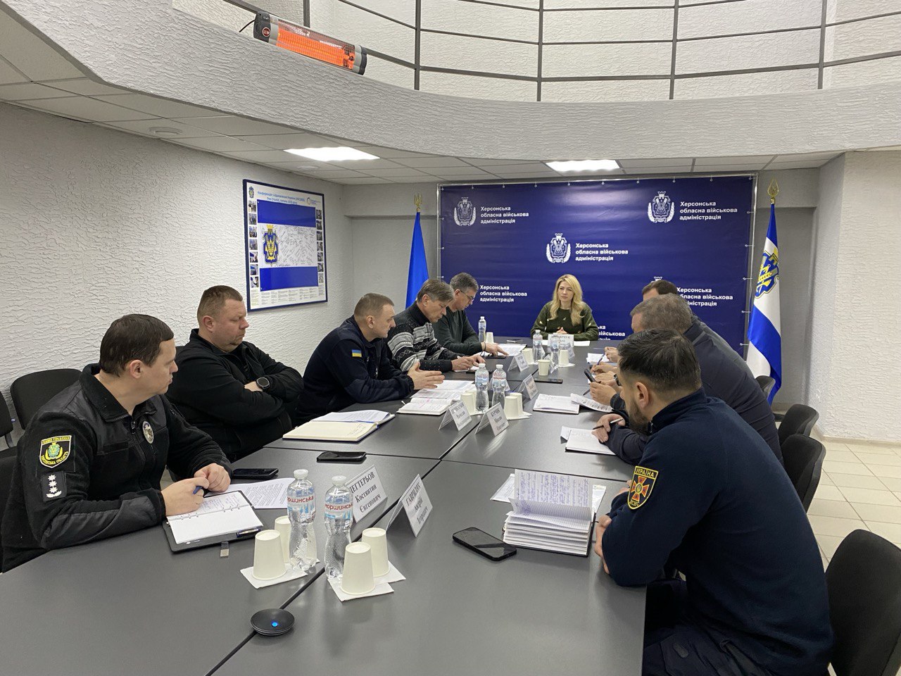Робоча нарада в Херсонській обласній військовій адміністрації. Група людей у цивільному та форменому одязі сидить за довгим столом у конференц-залі, обговорюючи документи. На фоні розташований синій банер із офіційною символікою адміністрації та державний прапор України.