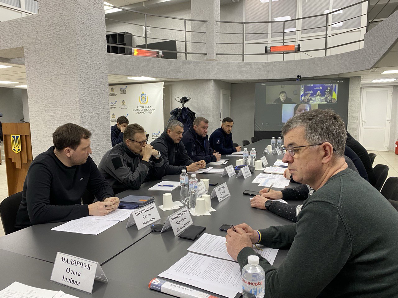 Учасники сидять за довгим столом, вивчають документи та ведуть обговорення. На столі встановлені таблички з іменами, стоїть вода в пляшках. На задньому плані видно великий екран для відеозв'язку та офіційну символіку обласної адміністрації.