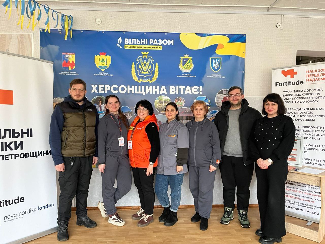 Сім представників команди (чоловіки та жінки) стоять у приміщенні на фоні великого синьо-жовтого банера гуманітарного центру «Вільні разом» (м. Кривий Ріг) з великим написом «Херсонщина вітає!» та гербами територіальних громад. По боках встановлені інформаційні стенди з логотипами організації Fortitude.