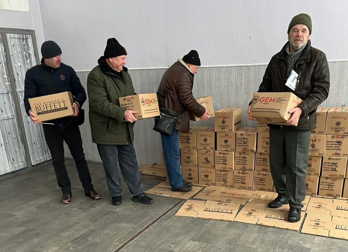 Чоловіки в зимовому одязі розвантажують та складають у приміщенні численні картонні коробки з гуманітарною допомогою. На коробках чітко видно логотипи благодійних організацій: The Howard G. Buffett Foundation та GEM (Global Empowerment Mission).