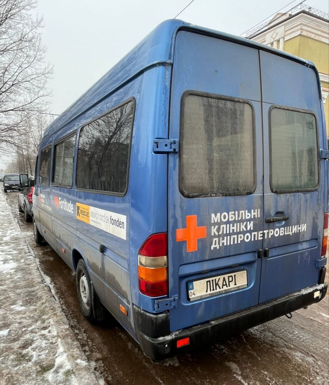 Синій мікроавтобус мобільної клініки Дніпропетровщини, брендований логотипами гуманітарних організацій, припаркований на засніженій узбіччі.