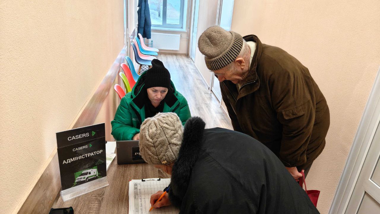 Адміністраторка медичної організації «CASERS» у зеленому фірмовому одязі консультує відвідувачів похилого віку за стійкою реєстрації в коридорі медичного закладу.