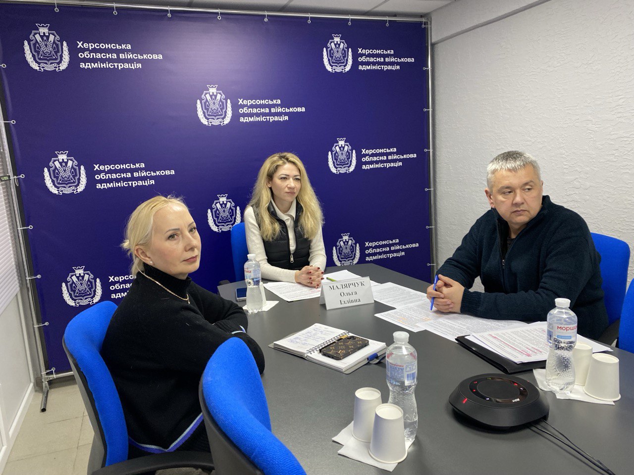Троє посадовців проводять робочу зустріч у кабінеті за столом із документами на фоні брендованого банера «Херсонська обласна військова адміністрація».