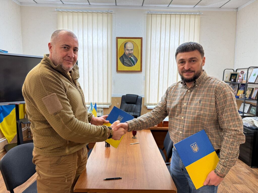 Двоє чоловіків у кабінеті потискають одне одному руки над робочим столом. Кожен із них тримає папку в кольорах національного прапора України з державним гербом. На стіні позаду них висить портрет Тараса Шевченка, а збоку стоїть прапор України.