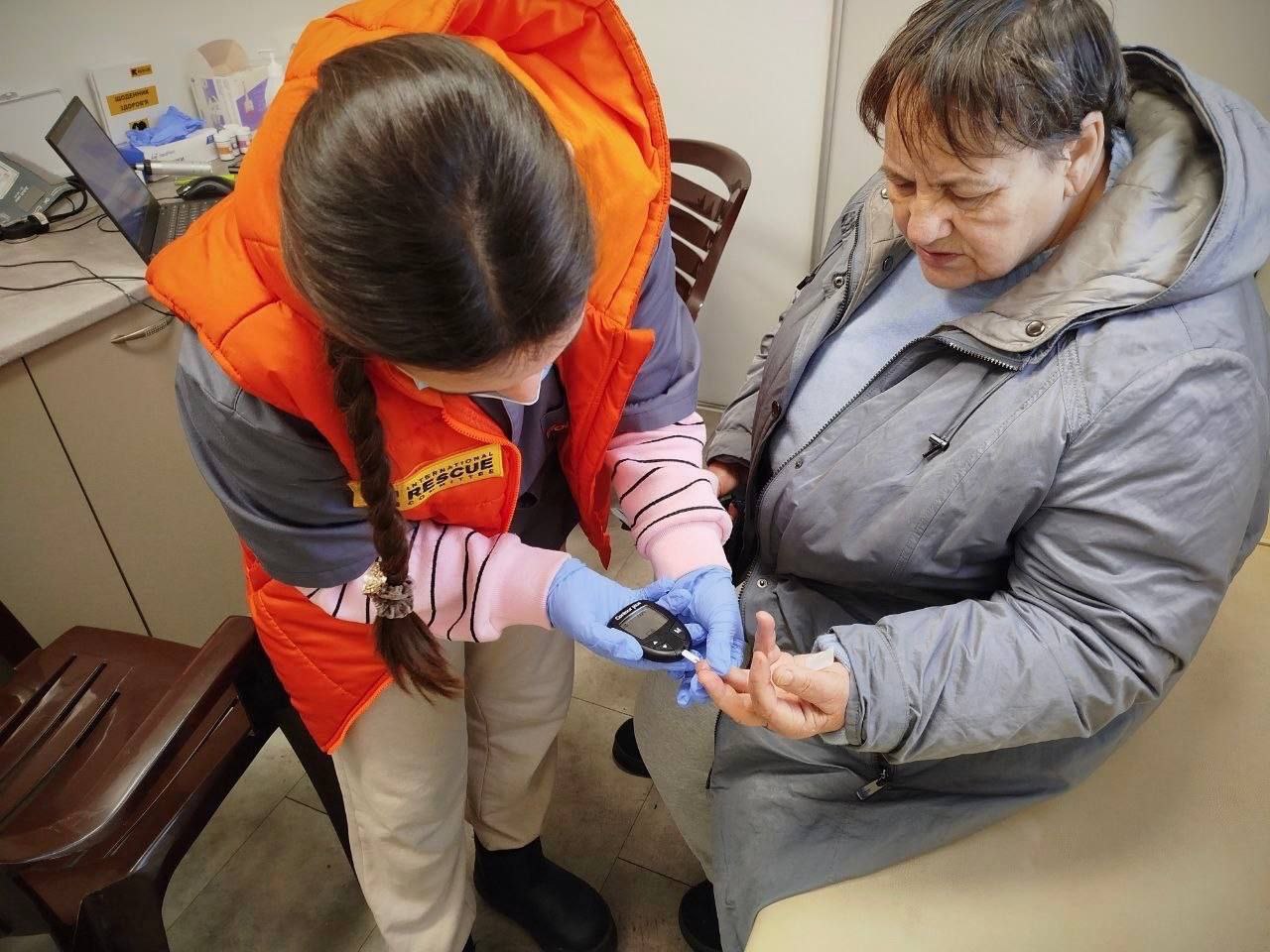 Медична працівниця в помаранчевому жилеті та рукавичках робить забір крові з пальця пацієнтки за допомогою глюкометра для перевірки рівня цукру.
