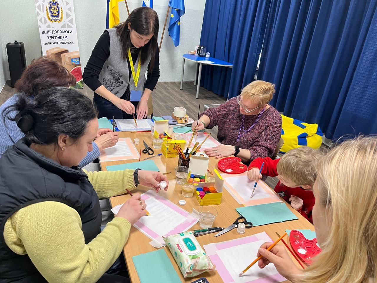 Група людей та дитина сидять за довгим столом у світлому приміщенні та займаються малюванням. На столі розкладені фарби, пензлі, палітри та аркуші паперу. На фоні видно банер «Центр допомоги херсонцям», український прапор та прапор Європейського Союзу.