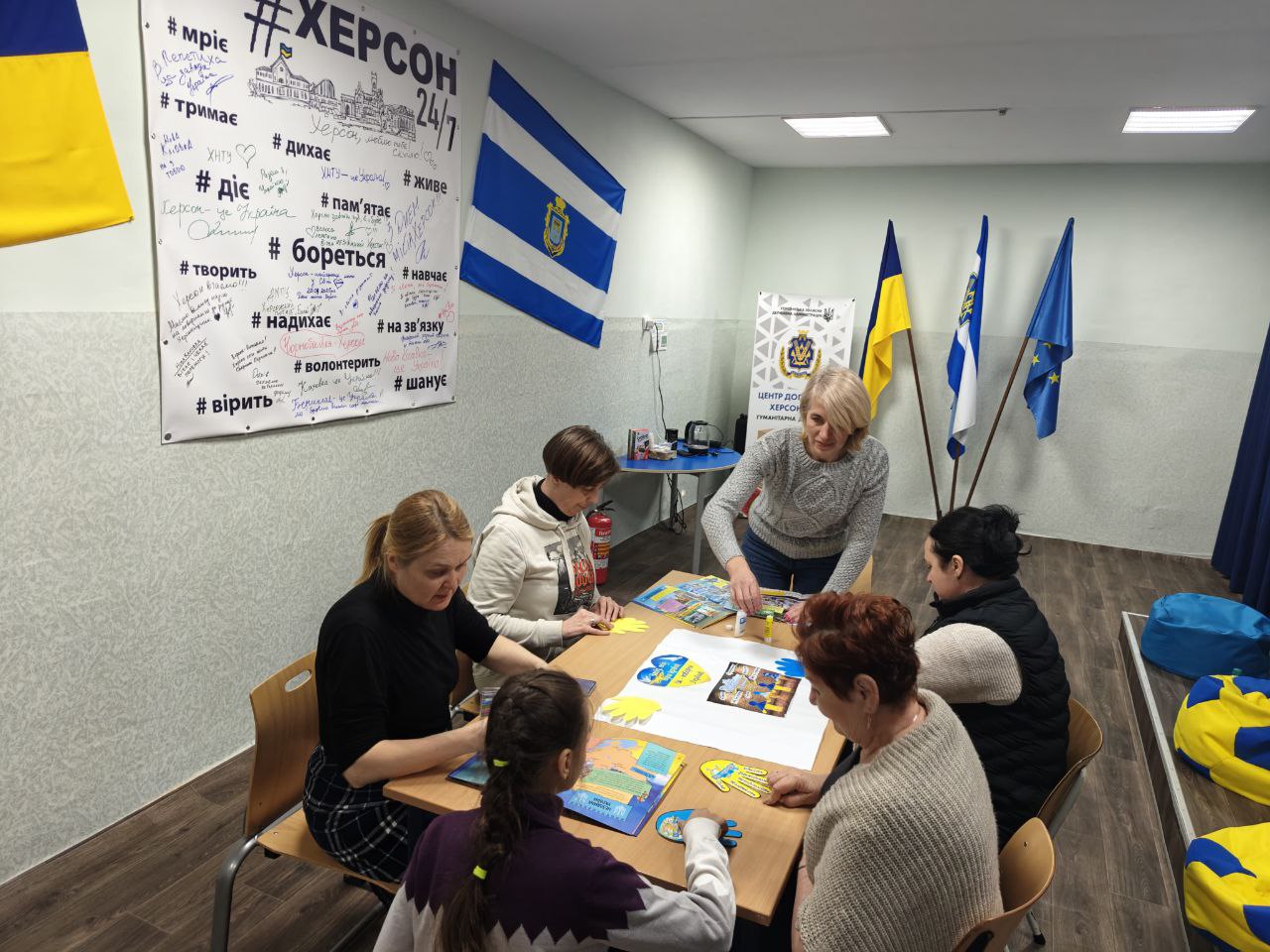Група людей різного віку сидить навколо столу в приміщенні з українською символікою та займається творчістю, створюючи спільний плакат із паперовими долоньками та малюнками. На стіні позаду — великий банер із написом «ХЕРСОН 24/7», патріотичними хештегами та побажаннями, а також прапори України, міста та ЄС.