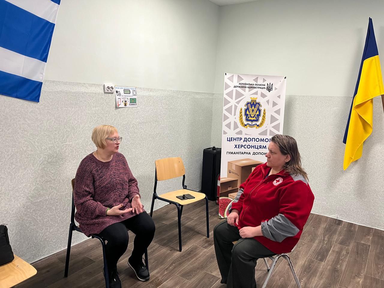 Дві жінки сидять на стільцях і спілкуються в офісному приміщенні. Поруч стоїть інформаційний банер із написом «Центр допомоги херсонцям», а на стінах розміщені прапори та інформаційні плакати.
