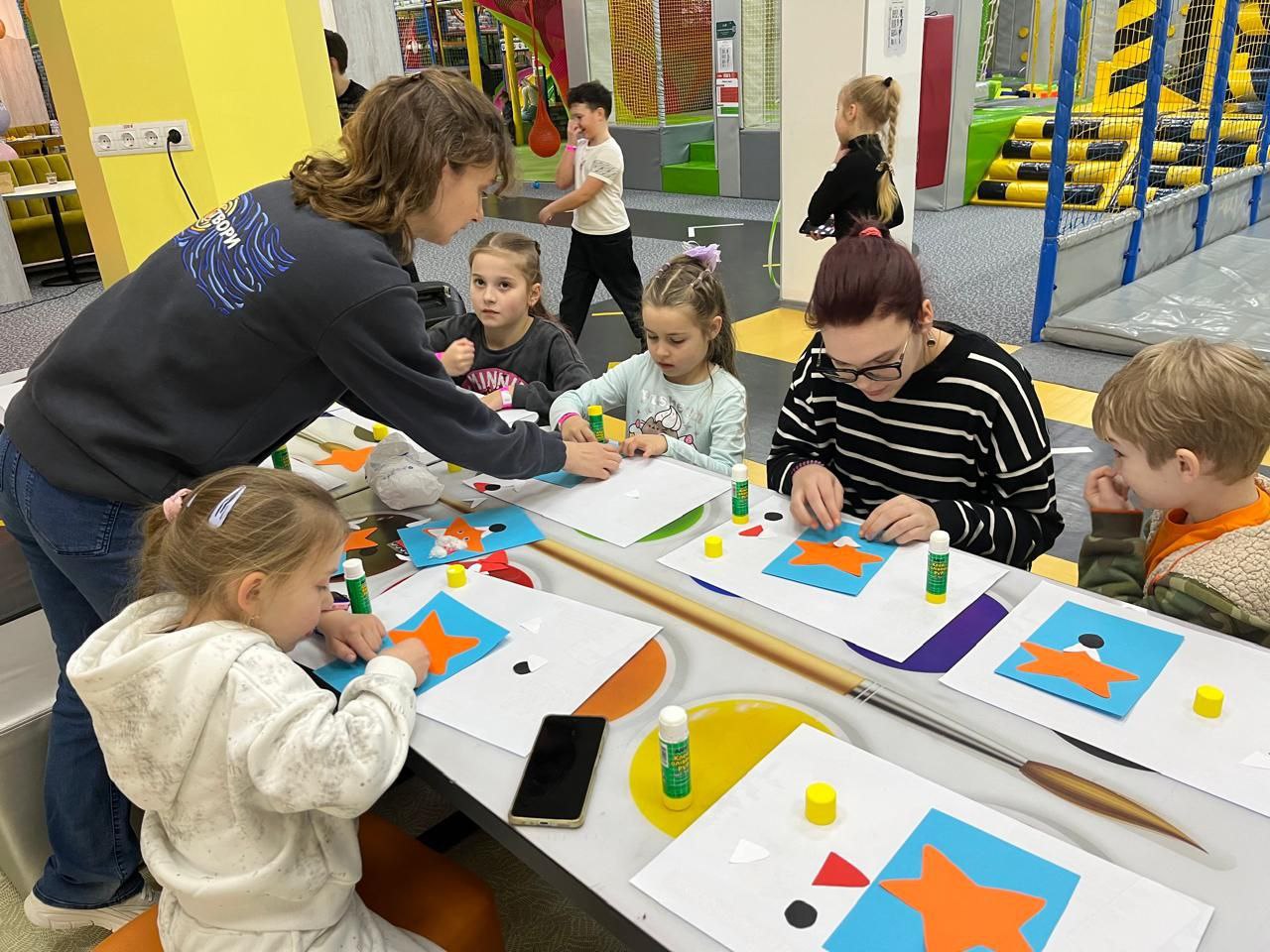 Діти та дорослі сидять за довгим столом у дитячому ігровому центрі, займаючись творчістю. Вони створюють аплікації з кольорового паперу за допомогою клею. На фоні видно ігрові конструкції та лабіринти.