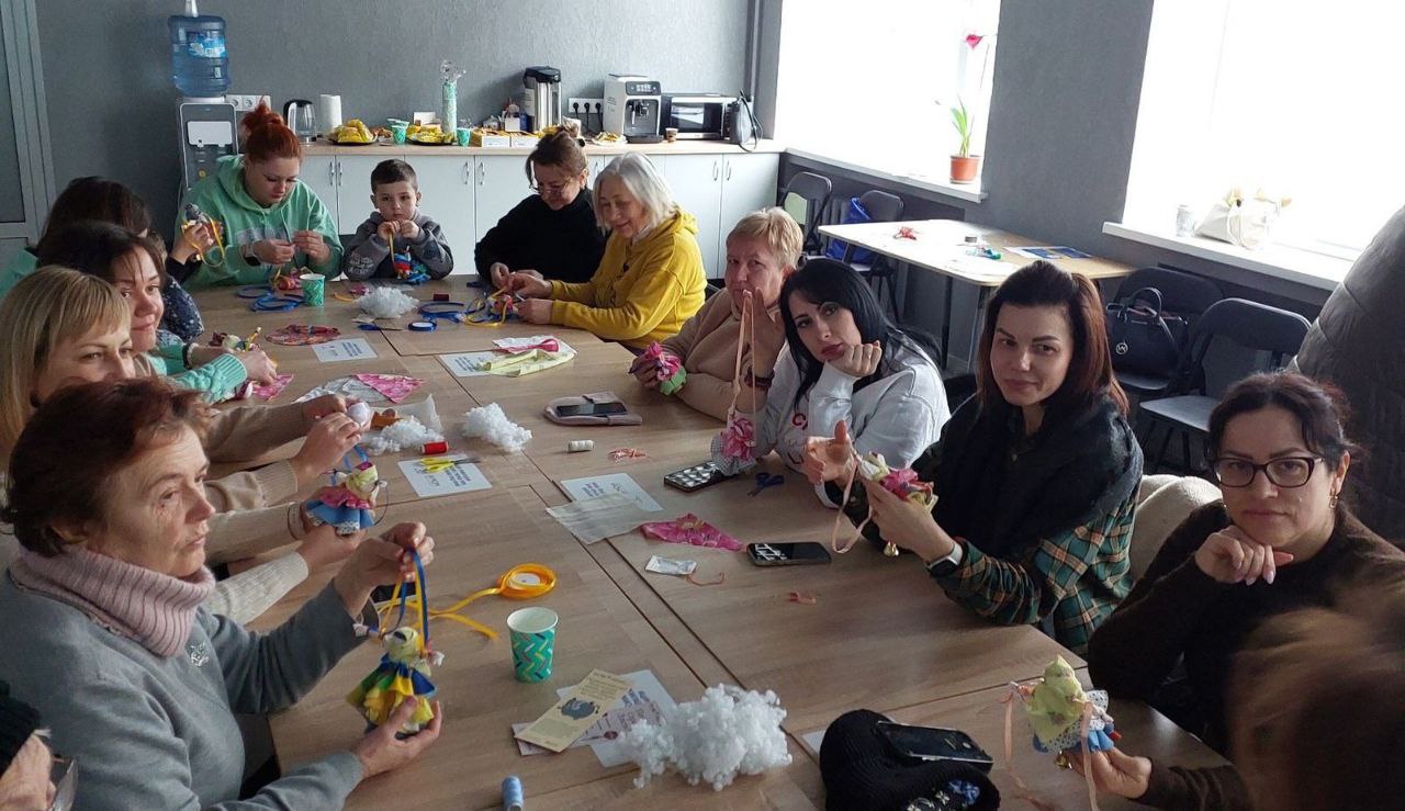 Група людей сидить за великим столом у світлому приміщенні та виготовляє традиційні ляльки з тканини. На столі лежать клаптики текстилю, стрічки та наповнювач. Учасники зосереджено працюють або демонструють свої вироби.