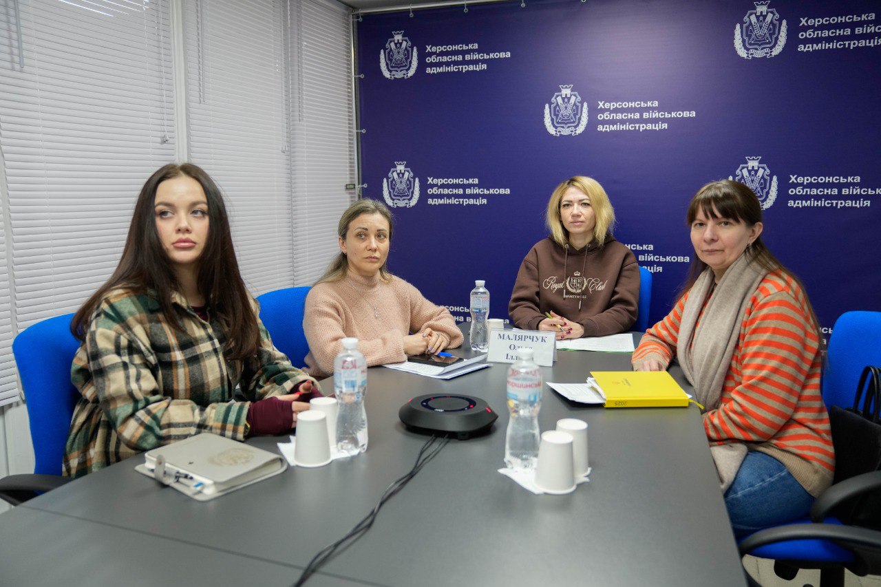 Чотири жінки сидять за робочим столом під час наради або офіційної зустрічі. На столі перед ними лежать блокноти, папери та стоять пляшки з водою. На задньому плані розташований фіолетовий банер із логотипом та написом «Херсонська обласна військова адміністрація».