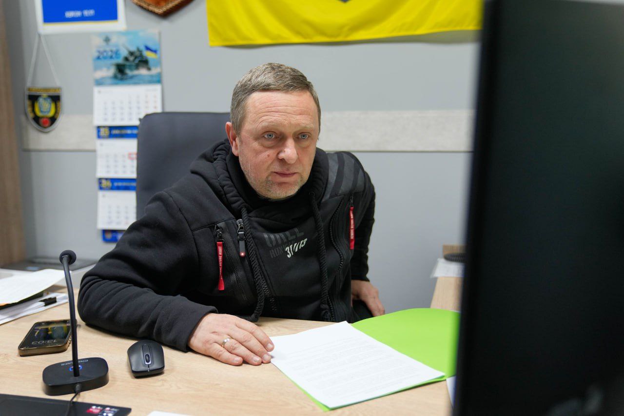 Чоловік у чорному худі сидить за робочим столом перед монітором комп'ютера. На столі лежать документи, смартфон та комп'ютерна миша, а на стіні позаду видно календар на 2026 рік та українську символіку.