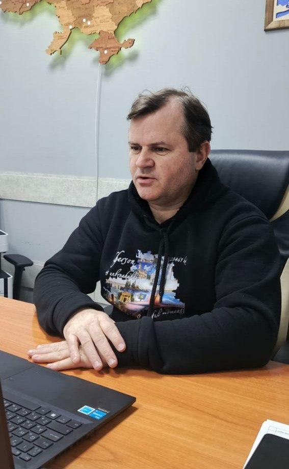 Чоловік сидить за дерев'яним столом, його руки складені перед ноутбуком, а на стіні позаду висить декоративна карта.