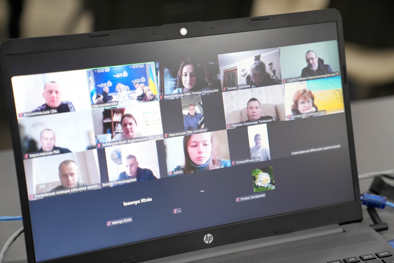 На екрані ноутбука відкрито вікно відеоконференції з багатьма учасниками. У верхній лівій плитці трансляції видно залу засідань із прапором України та логотипом Херсонської ОВА.