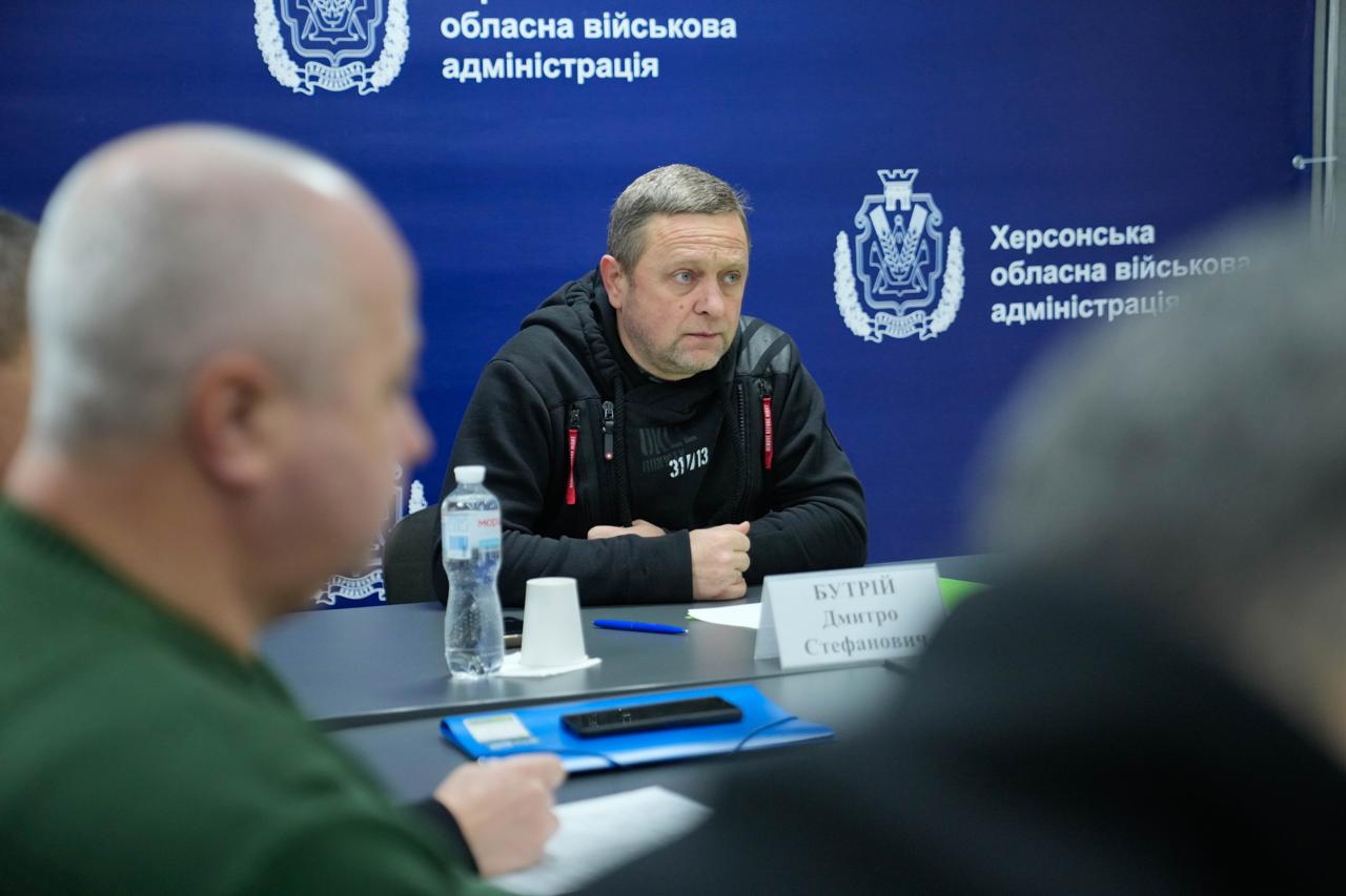 Портретне фото чоловіка за столом під час наради. Він одягнений у чорну кофту з капюшоном, руки складені перед собою. На фоні — синій банер із символікою Херсонської обласної військової адміністрації.