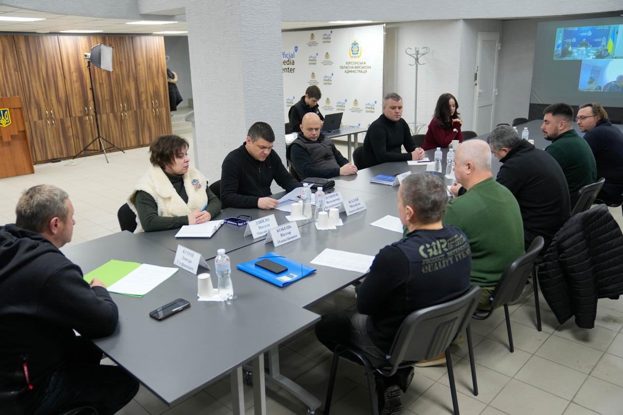 Робоча нарада представників місцевої влади у конференц-залі. Учасники сидять за столом з документами, на фоні розміщено банери «Медіацентр Україна» та Херсонської ОВА. У кутку зали видно екран із трансляцією відеоконференції.