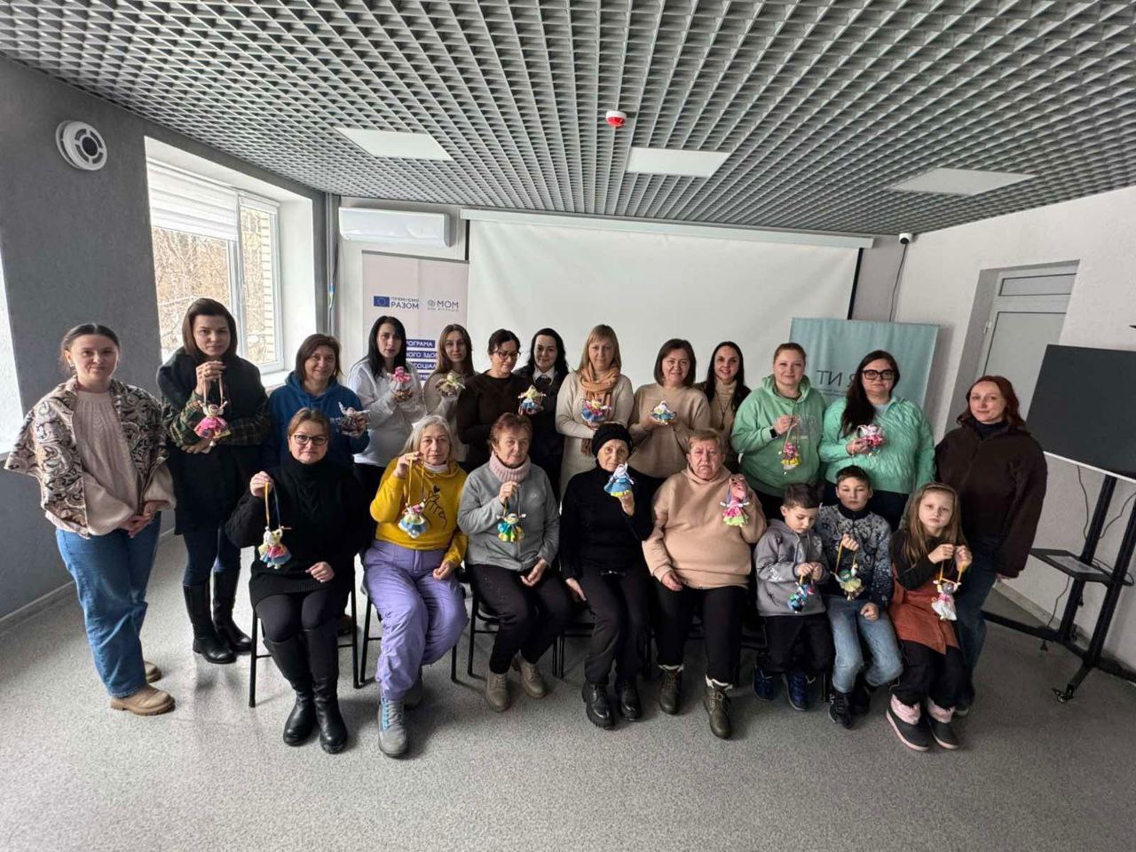 Група людей різного віку позує в приміщенні, тримаючи в руках саморобні ляльки-мотанки. Вони стоять і сидять на тлі білого екрана та банерів з логотипами організацій.