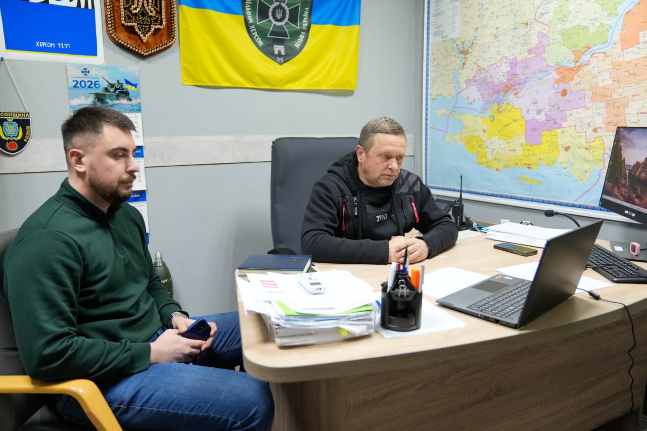 Двоє чоловіків у кабінеті беруть участь в онлайн-конференції через ноутбук. На стіні за ними розміщено карту області та прапор. На столі лежать документи та канцелярське приладдя.