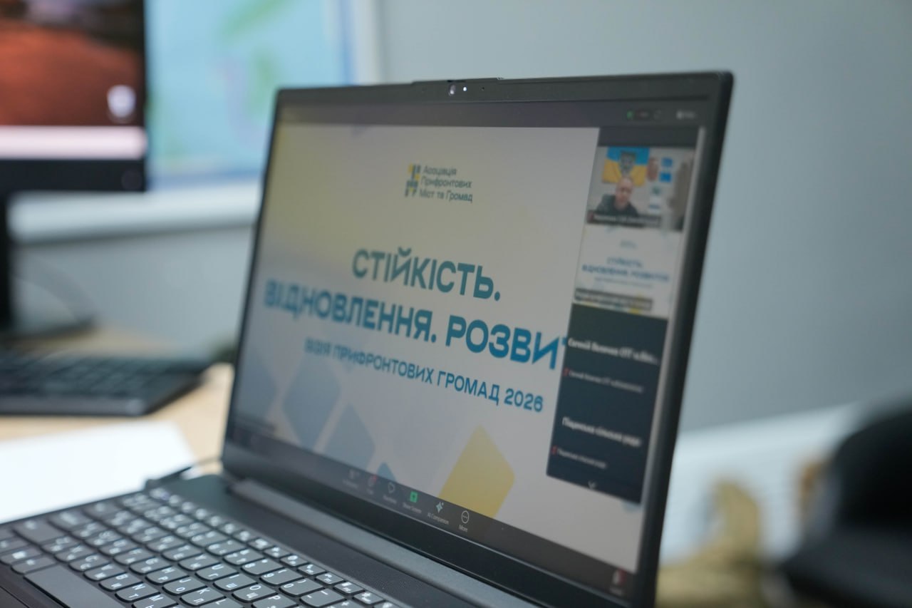 Ноутбук на робочому столі, на екрані якого відкрито онлайн-конференцію. Видно головний слайд із написом «СТІЙКІСТЬ. ВІДНОВЛЕННЯ. РОЗВИТОК ПРИФРОНТОВИХ ГРОМАД 2026» та логотип Асоціації прифронтових міст та громад.