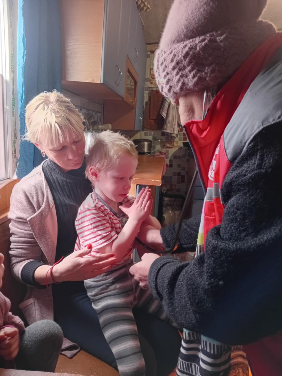 Працівник гуманітарної місії у фірмовому жилеті проводить медичний огляд дитини за допомогою стетоскопа в домашніх умовах. Мати підтримує дитину під час процедури.