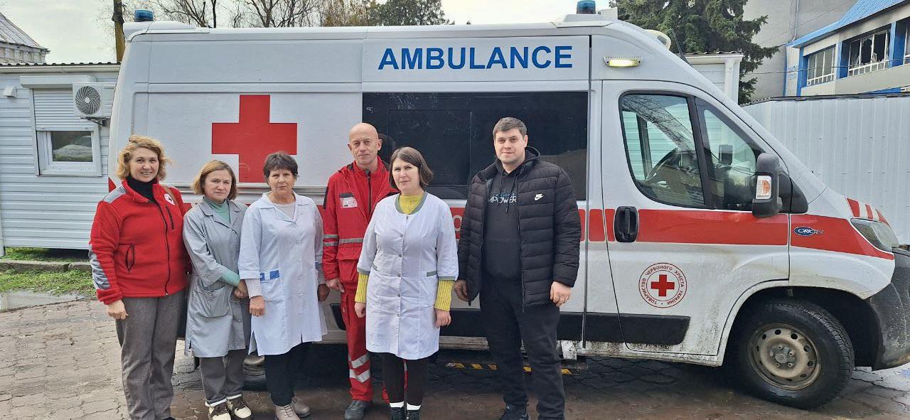 Група медичних працівників та волонтерів стоїть на фоні автомобіля швидкої допомоги з написом «AMBULANCE» та емблемою Червоного Хреста. Люди одягнені в медичні халати та спецодяг, позуючи для спільного фото на вулиці.