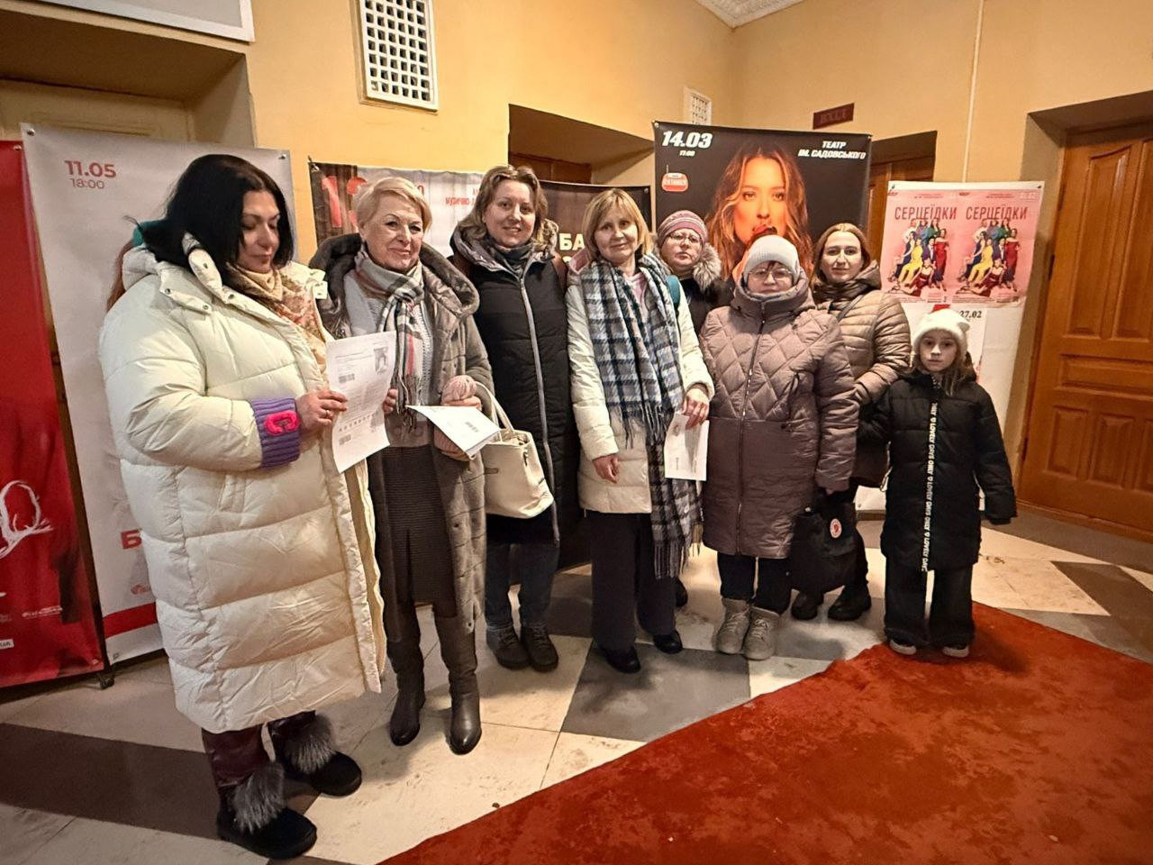 Група жінок та дитина стоять у фоє театру на фоні афіш. Дехто з присутніх тримає в руках паперові квитки або документи.