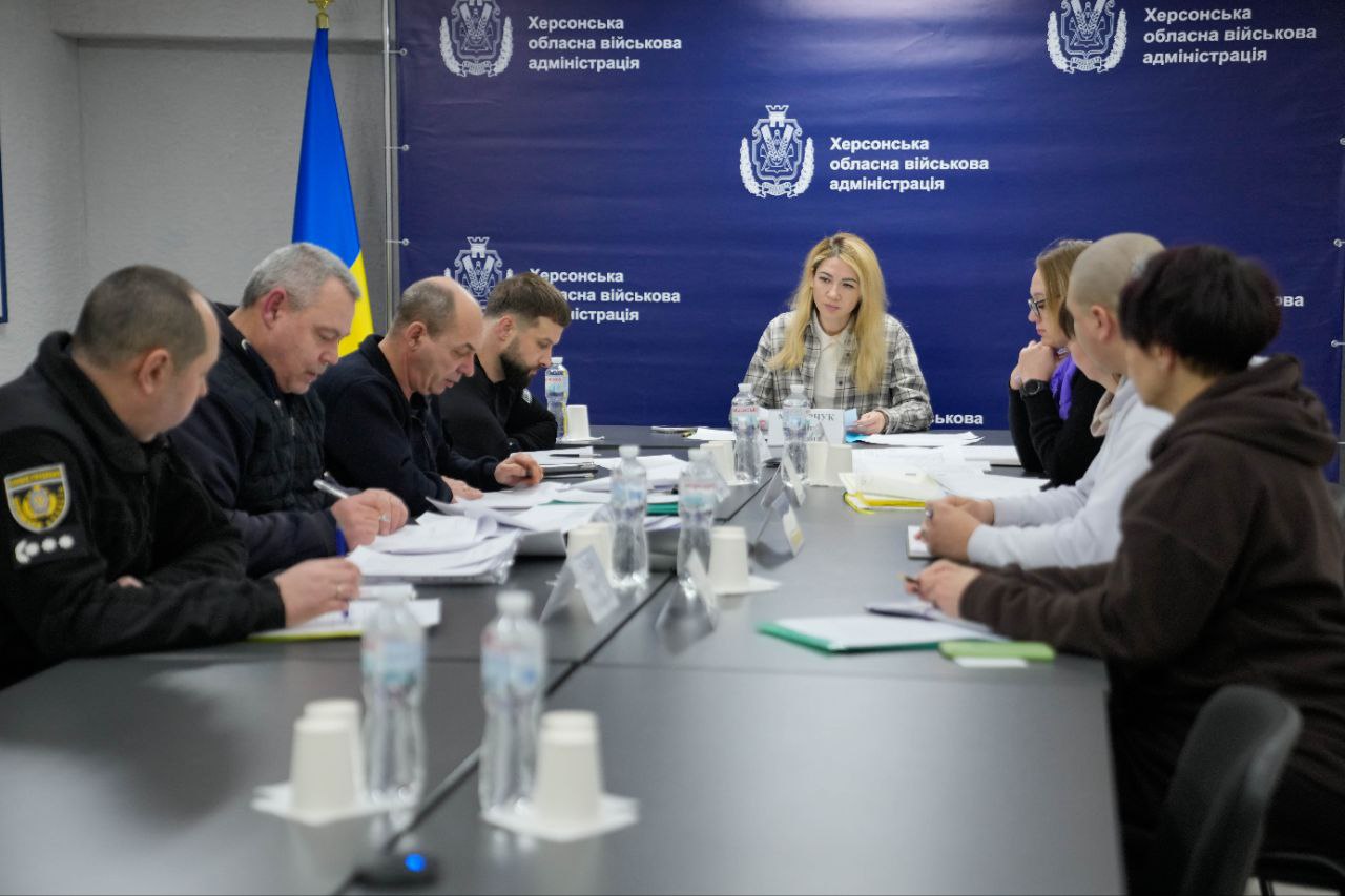 Група людей проводить робочу зустріч за великим столом. Учасники працюють з документами на фоні стіни з логотипами обласної військової адміністрації.