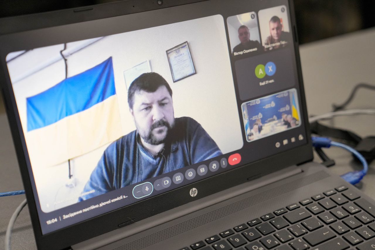 На екрані ноутбука відображено інтерфейс відеодзвінка. У головному вікні — чоловік на тлі прапора України та дипломів на стіні. У бічній панелі видно інших учасників зустрічі та іконки налаштувань зв'язку.