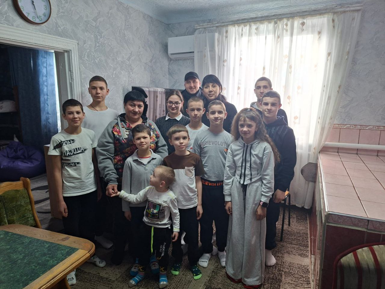 Групова фотографія дорослих та дітей різного віку, які позують у світлій кімнаті біля вікна. Серед присутніх у центрі — представник поліції у формі.