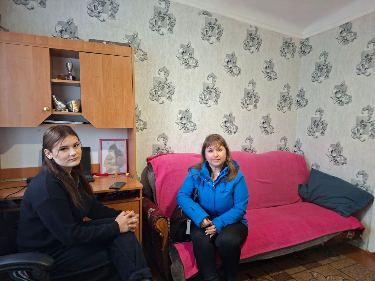 Дві жінки сидять у кімнаті та спілкуються. Одна з них сидить за робочим столом, інша — на дивані в синій куртці.