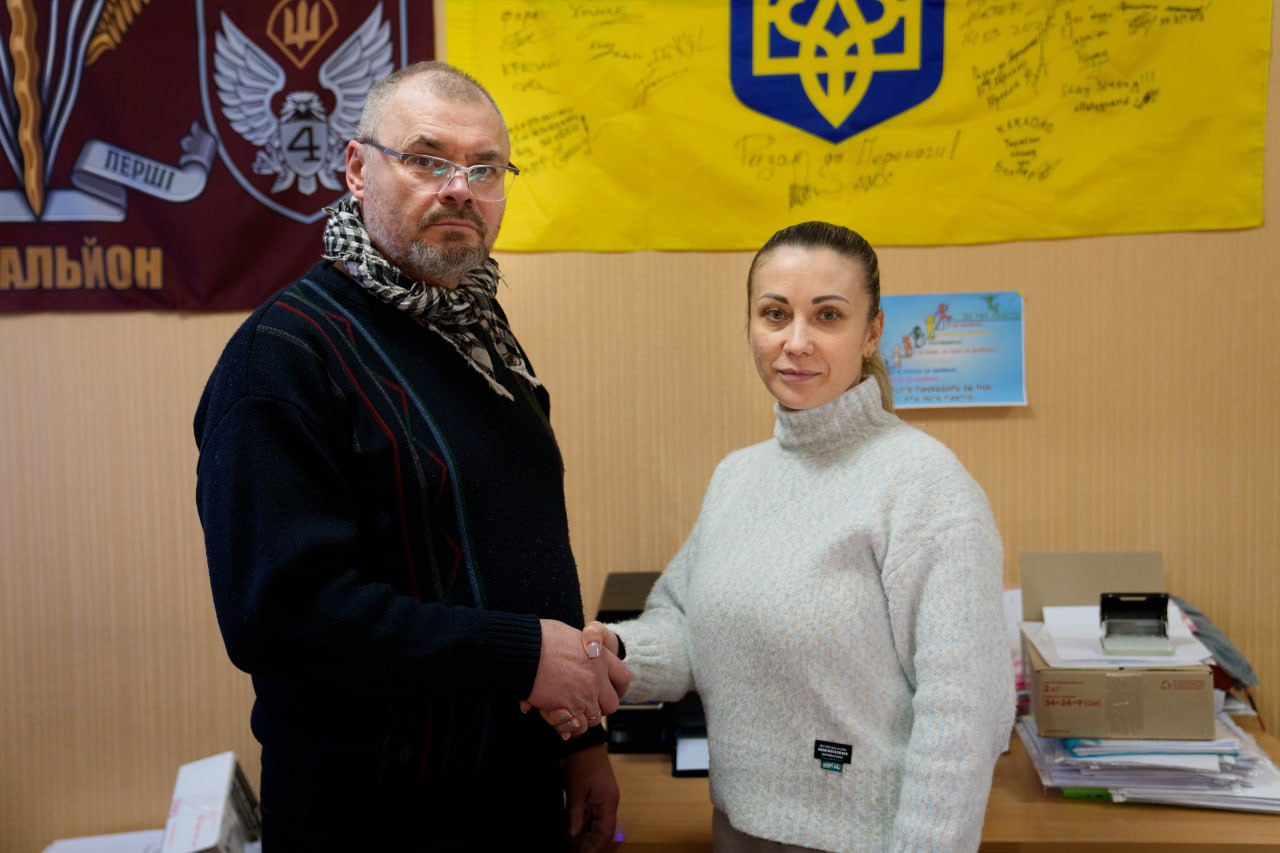 Чоловік та жінка тиснуть один одному руки, стоячи в офісному приміщенні. На стіні позаду них висять прапори: жовто-блакитний із підписами та військовий стяг із символікою батальйону. На передньому плані видно робочий стіл із документами та канцелярським приладдям.