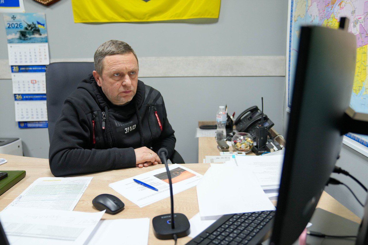 Чоловік у чорному худі сидить за робочим столом, зосереджено дивлячись на монітор комп'ютера. На столі лежать документи, мікрофон та робочі записи. На стіні позаду нього — карта, календар на 2026 рік та державний прапор.