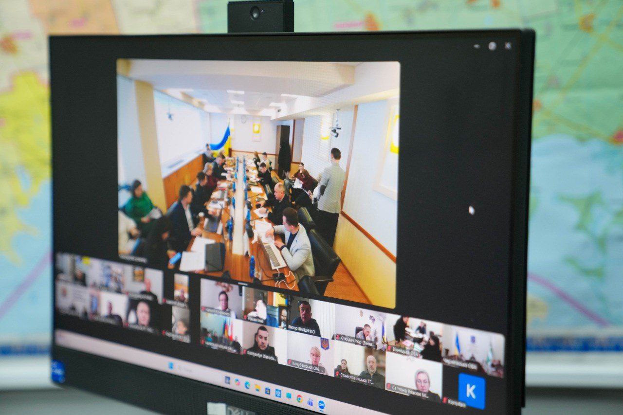 Монітор комп'ютера, на якому відображено вікно відеоконференції з багатьма учасниками. У центральній частині екрана видно великий зал засідань з людьми за столом, а внизу — мініатюри з відео окремих учасників наради. На фоні за монітором розміщена географічна карта.