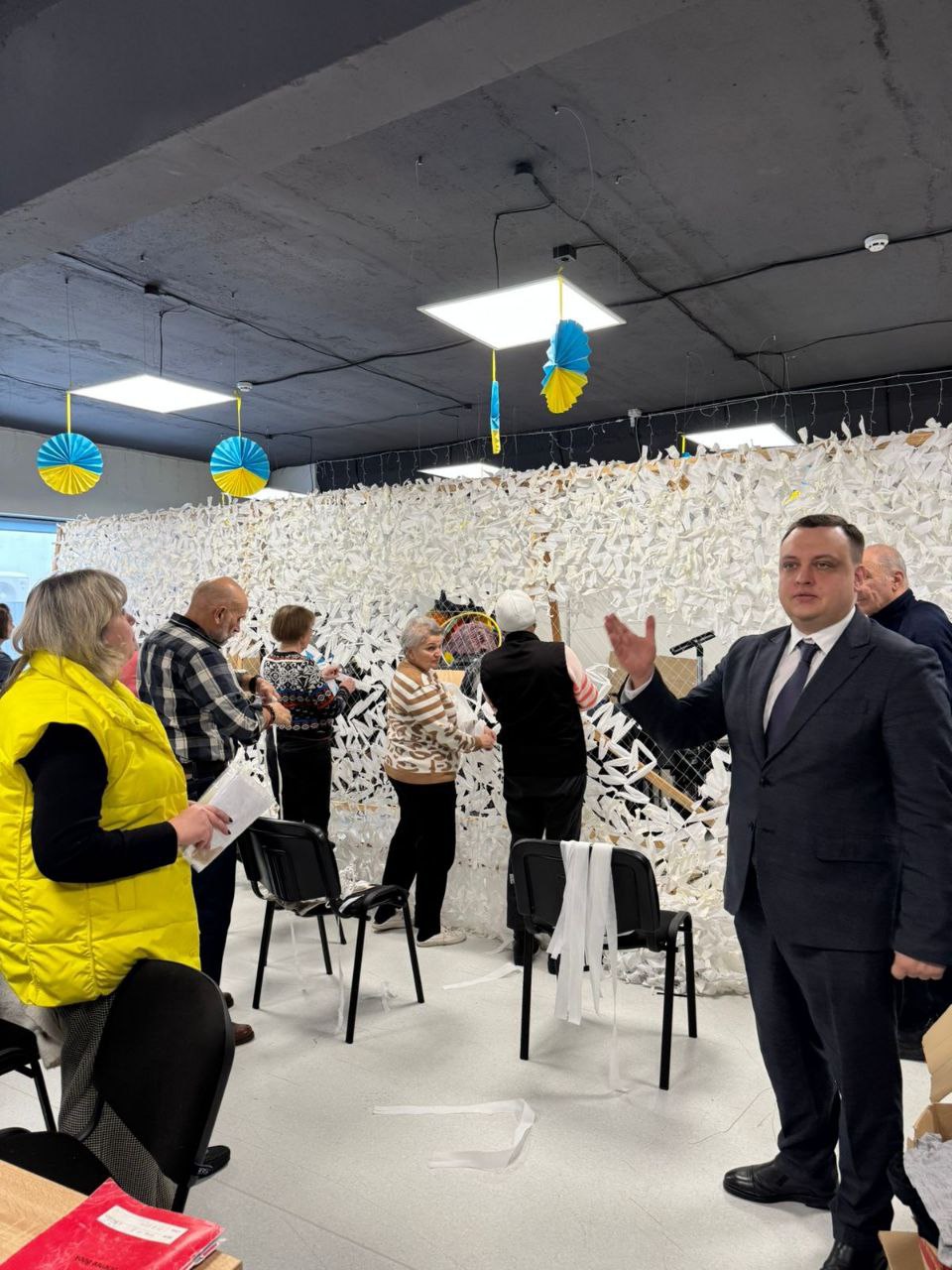 Група людей у просторому приміщенні займається плетінням білої маскувальної сітки. На передньому плані чоловік у діловому костюмі спілкується з присутніми. Стеля декорована синьо-жовтими паперовими прикрасами.