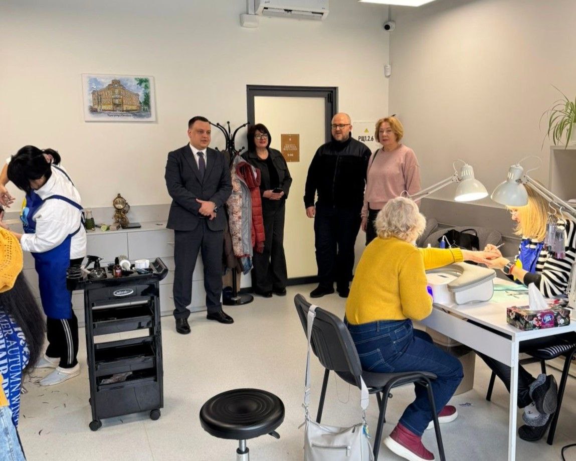 Група людей в офіційному та повсякденному одязі відвідує салон надання побутових послуг. У приміщенні видно робочі місця для манікюру та перукарських послуг, де майстри працюють із клієнтами.