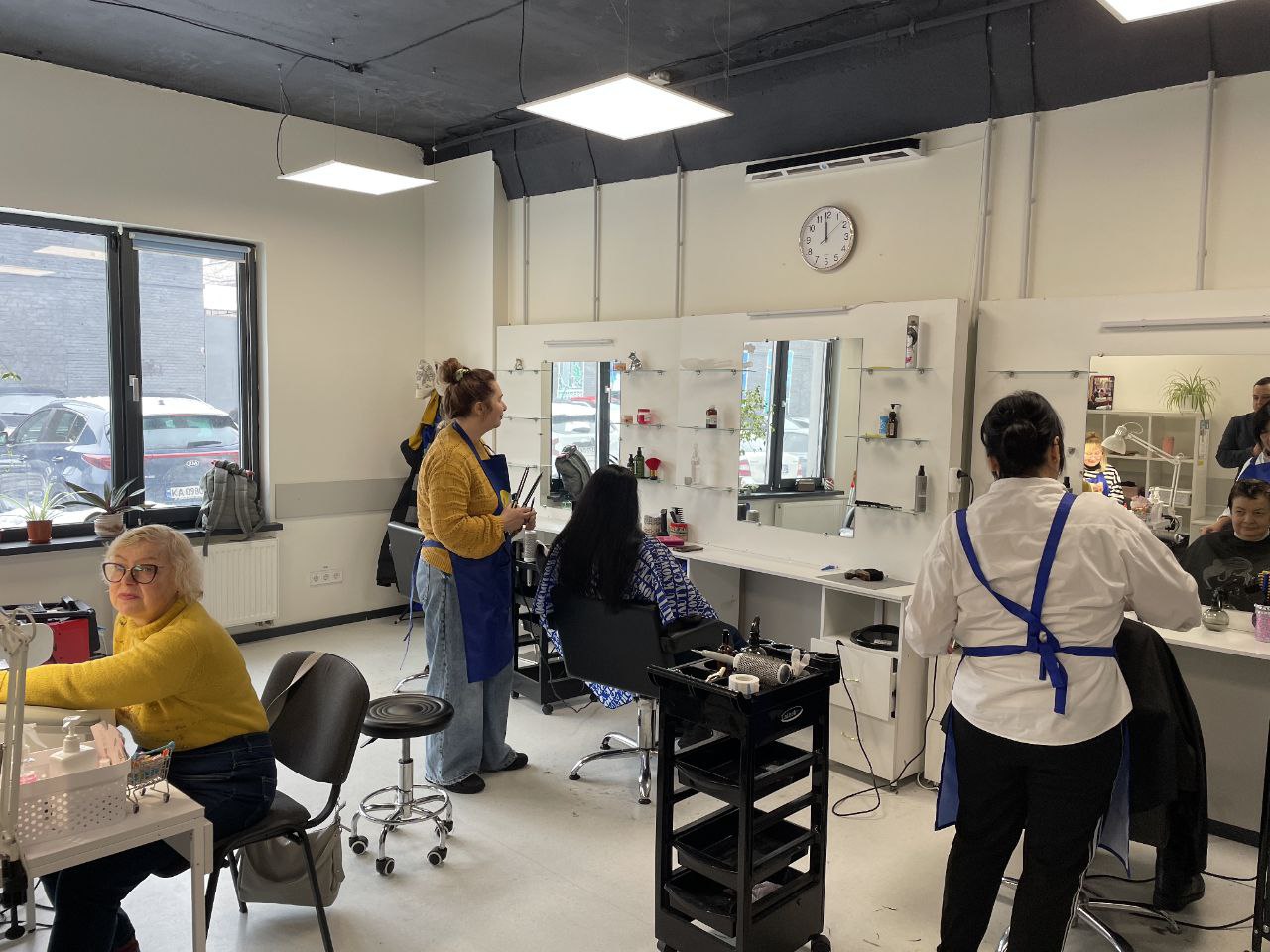 Інтер'єр соціальної перукарні, де майстри у синіх фартухах надають послуги клієнтам. У приміщенні облаштовані робочі місця з великими дзеркалами, полицями для інструментів та професійними кріслами. Крізь вікно видно припарковані на вулиці автомобілі.